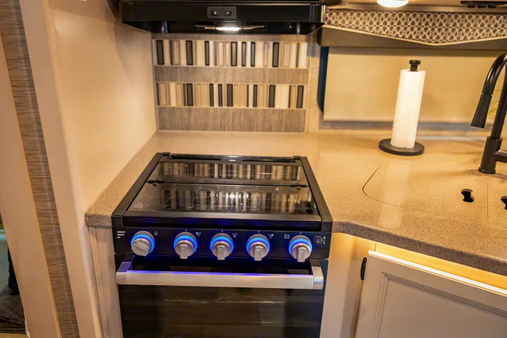 Modern RV rental kitchen features a black stove with glowing blue knobs, speckled countertop, and sink area with a black faucet.. Thor A.C.E. 2023