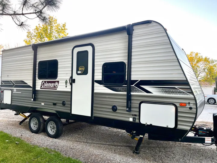 A modern grey and white Coleman travel trailer, perfect for an RV rental, stands ready on a gravel lot.. Dutchmen Coleman Lantern 2023