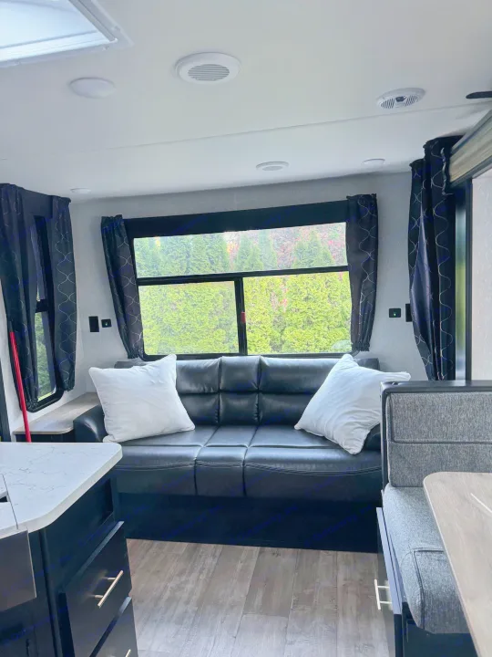Inside a modern RV rental: a black leather sofa with white pillows, large windows, and dinette. Cozy travel living space.. Greywolf 23MK 2022