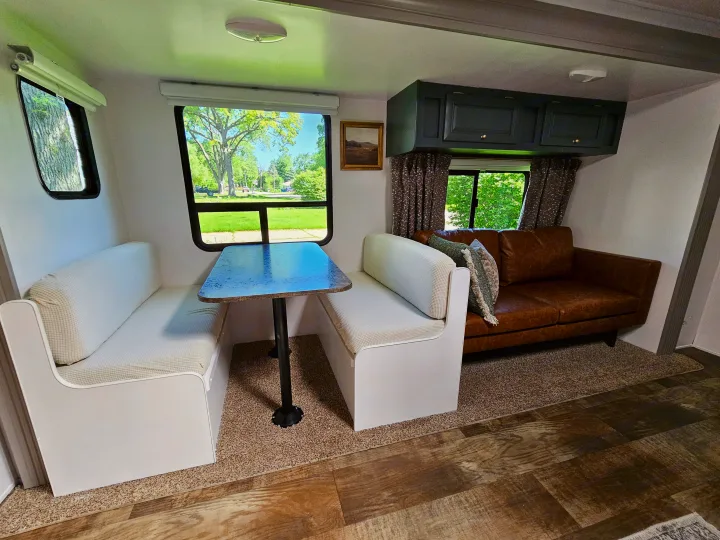 Bright RV rental interior showing a comfortable dining booth & brown sofa, with large windows overlooking green scenery.. Crossroads RV Z-1 2017