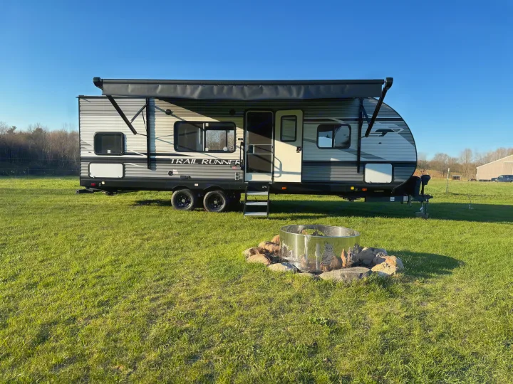Modern travel trailer with extended awning and fire pit on green grass, perfect for your next RV rental.. Heartland RVs Trail Runner 2023