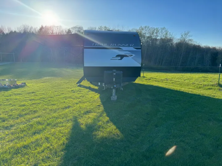 Sunny field scene: A 'Trail Runner' RV rental with extended awning and fire pit awaits, backed by trees.. Heartland RVs Trail Runner 2023