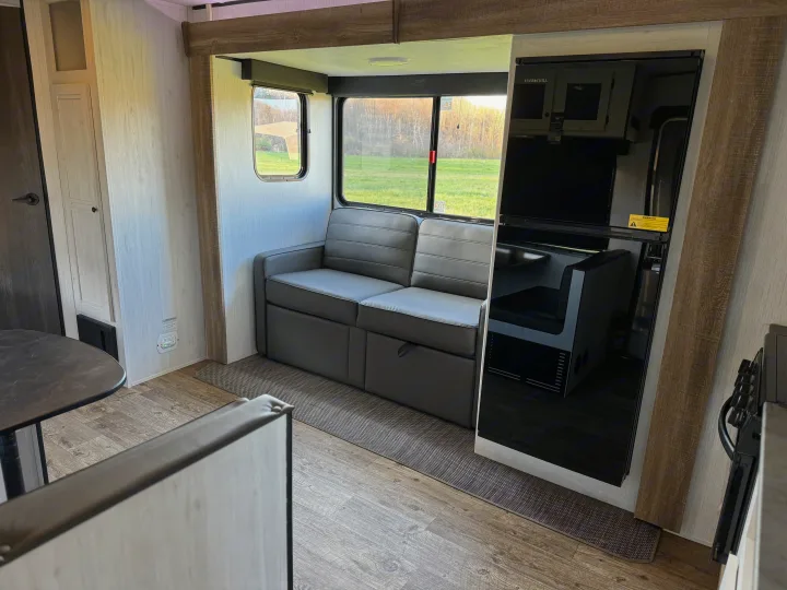 Interior of an RV rental showing a gray sofa, large windows with a field view, and a reflective screen showing a dining booth.. Heartland RVs Trail Runner 2023