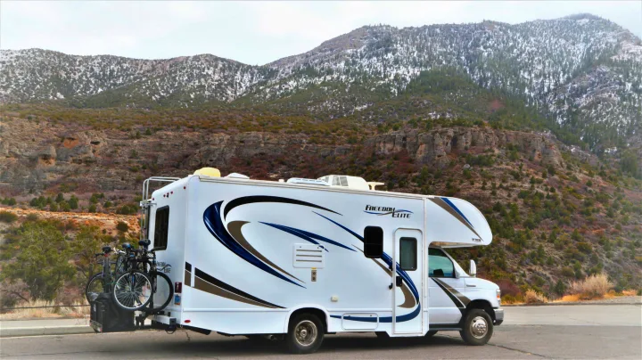 A white Class C **RV rental**, with bikes on the back, is parked on a mountain road with snow-dusted peaks behind it.. Thor Motor Coach Freedom Elite 2018