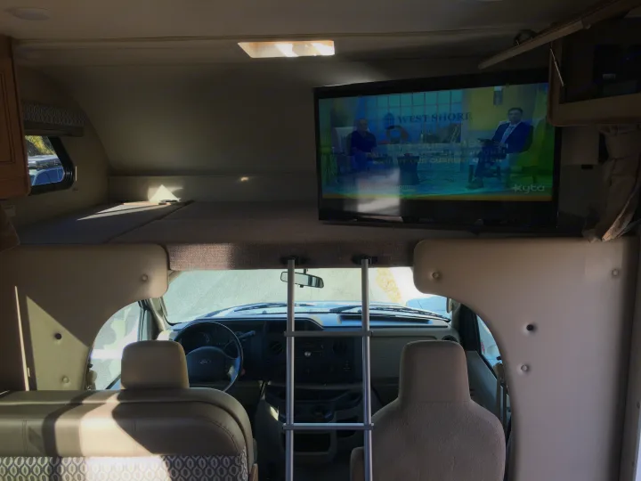 Inside an RV rental, a TV plays above the driver's cab, with an overhead bunk and ladder visible.. Thor Motor Coach Freedom Elite 2018