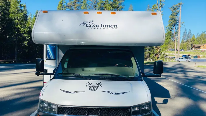 Front view of a white Coachmen Freelander RV rental with an Autobot symbol on the hood, parked outdoors among trees.. Coachmam Freelander 21QB 2012