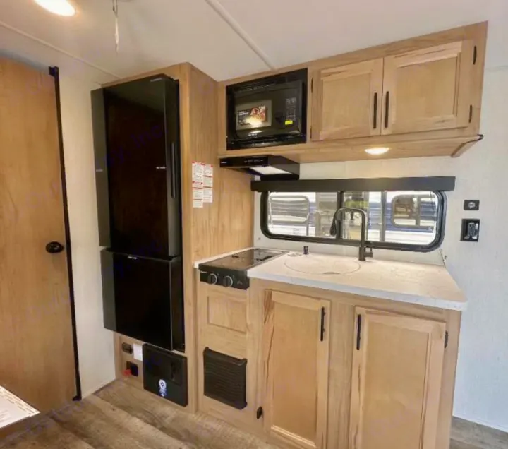 RV rental kitchen: a tall black fridge, microwave, 2-burner stove, sink, and light wood cabinets, ready for your trip.. Forest River Cherokee Wolf Pup 2023