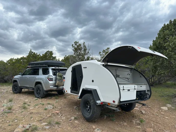 A silver SUV with a rooftop tent tows a white teardrop RV rental, parked off-road for adventure camping under cloudy skies.. Escapod Topo 2 - Nomad 2024