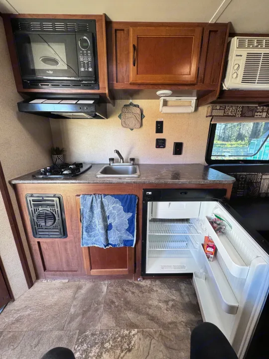 This rv rental's compact kitchen features a microwave, two-burner stove, sink, and open fridge, all neatly arranged.. Jayco Jay Flight SLX 2016