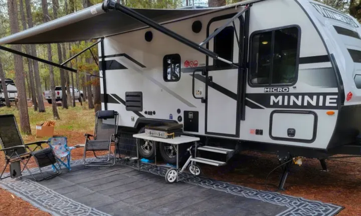 This RV rental features a white Micro Minnie travel trailer with an extended awning, chairs, and grill set up in a wooded campsite.. Minnie Micro Minnie 2023