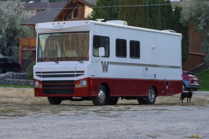 White & maroon Winnebago Brave RV rental, parked on a gravel lot with a dog. Your adventure begins!. Winnebago Brave 2015