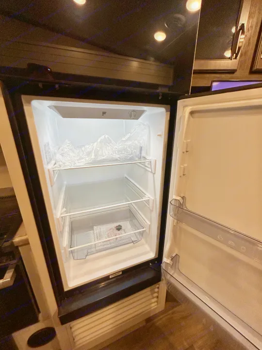 An open, new refrigerator in an RV rental, showing clean shelves, plastic wrap, and a crisper drawer.. Keystone RV Cougar 2023