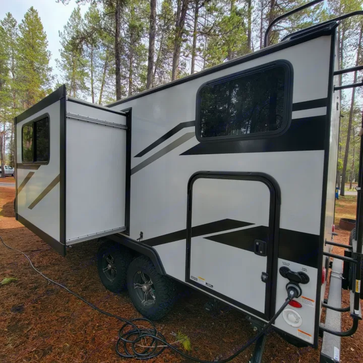 Modern white travel trailer with slide-out extended, parked at a wooded campsite with power hookup. Great RV rental option!. Minnie Micro Minnie 2023