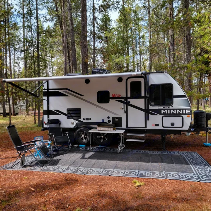 This Micro Minnie RV rental is perfectly set up at a wooded campsite with an extended awning, chairs, grill, and outdoor mat.. Minnie Micro Minnie 2023