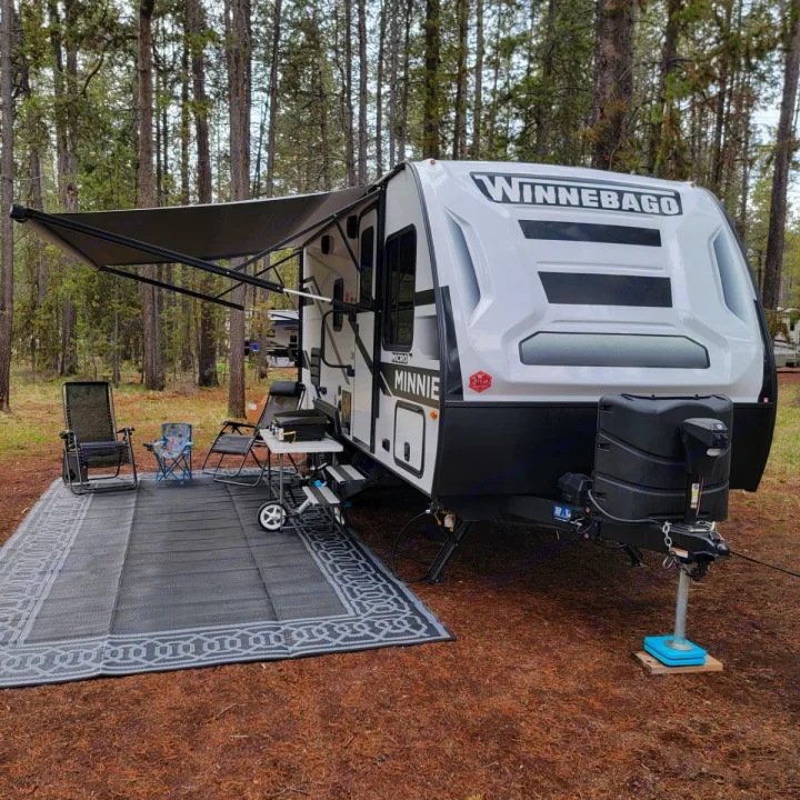 Winnebago Minnie Micro RV rental at a forest campsite with awning, chairs, and outdoor rug for comfortable living.. Minnie Micro Minnie 2023