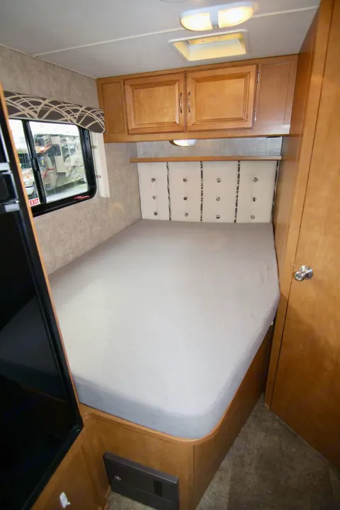 Interior view of an RV rental bedroom showing a bed with light gray sheet, wooden cabinets, and a window.. Winnebago Minnie Winnie 2016