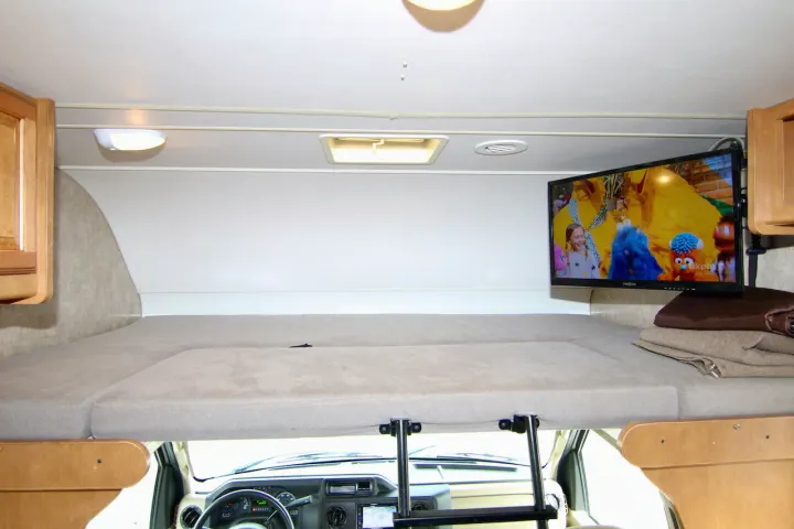 Inside an RV rental: a cozy cab-over bunk with a bed, TV playing cartoons, and a ladder leading to the driver's area.. Winnebago Minnie Winnie 2016