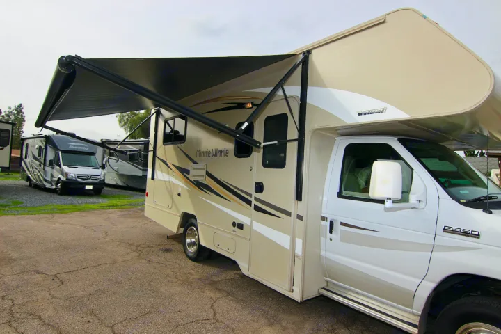 A beige and white Winnebago Minnie Winnie Class C RV with its black awning extended, parked in a lot, ideal for rv rental.. Winnebago Minnie Winnie 2016