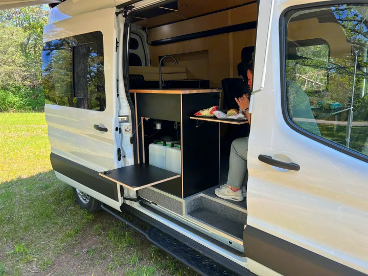 Enjoying a meal inside a custom RV rental. Features a clever interior kitchen with fold-out tables & water tanks for mobile living.. Ford Transit 2023