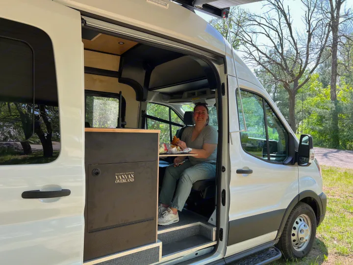 A smiling woman enjoys her burger inside a white Vanvan Overland RV rental, ready for travel.. Ford Transit 2023