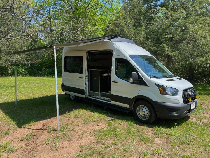 White camper van RV rental parked in a sunny, grassy clearing with its awning extended, perfect for outdoor adventures.. Ford Transit 2023