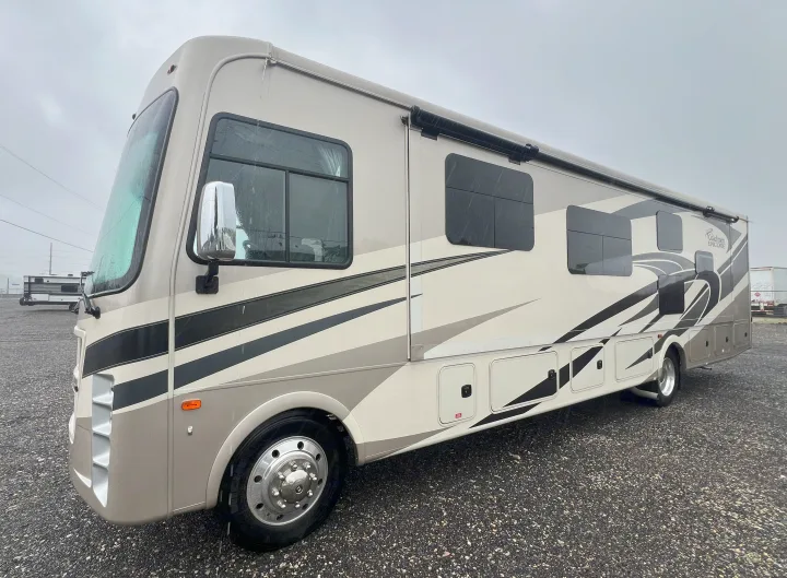 Experience comfort with this Class A Coachmen Encore RV rental. Beige motorhome on gravel under an overcast sky.. Coachmen Encore 2021