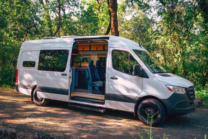 White camper van RV rental, side door open to a custom interior, parked on a dirt path in a sunny green forest.. Mercedes-Benz Sprinter 2024