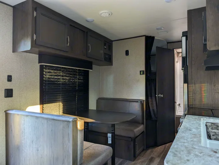 RV rental interior: a modern dinette with dark cushions & table, overhead cabinets, kitchen counter, and bathroom entrance.. Jayco Jay Flight SLX 2021