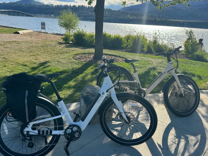 Two white OKAI e-bikes parked by a sunny lake with mountains & a bridge, ideal for exploring after an RV rental adventure.. Ebike 500 Watt 2024