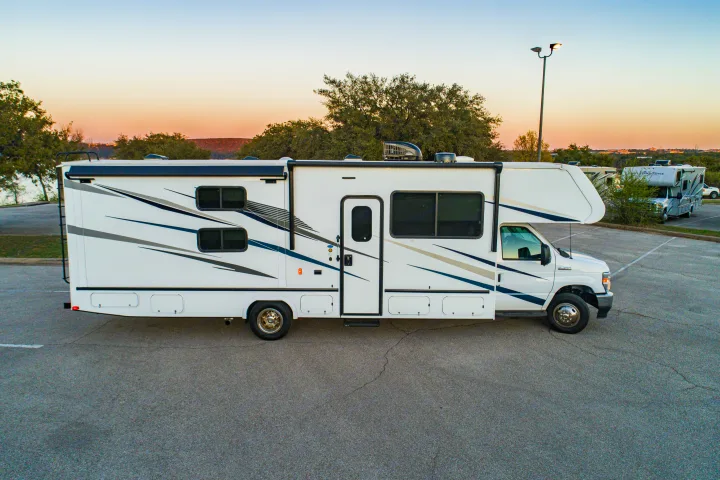Side view of a modern white RV rental with black/blue accents, parked on asphalt at sunset. Trees and light pole visible.. Gulf Stream Yellowstone 2023