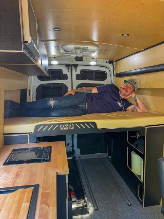Inside an RV rental, a man relaxes in a custom camper van with a cozy bed & kitchen.. Ford Transit 2023