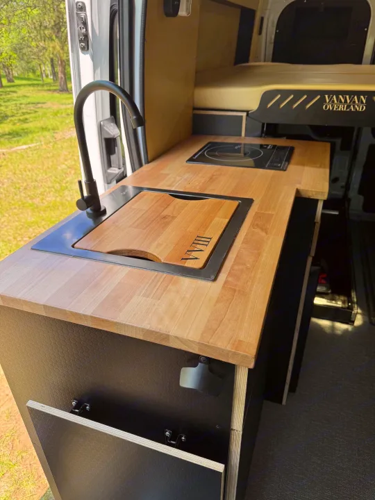 RV rental interior: A compact kitchen with a light wood countertop, black sink, and induction cooktop. A bed is visible in the background.. Ford Transit 2023