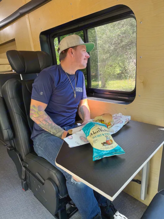 A man with tattoos enjoys a sandwich and chips inside a cozy RV rental, looking out a window at nature.. Ford Transit 2023
