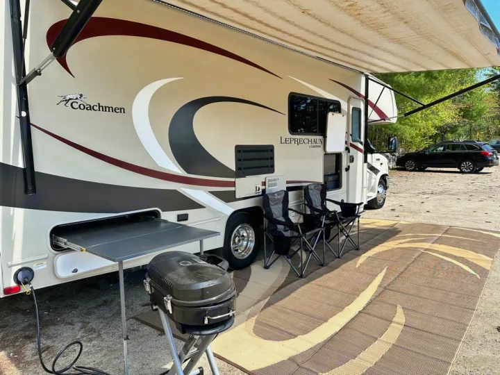 Enjoying an RV rental adventure: a spacious motorhome set up with an awning, chairs, and grill at a serene campsite.. Coachmen Leprechaun 2019
