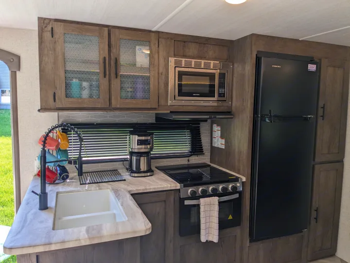 Inside an rv rental: this modern, compact RV kitchen. Features a black refrigerator, microwave, oven, sink, and coffee maker.. Forest River Wildwood 2021