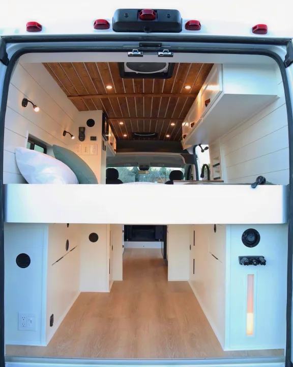 Spacious & modern camper van interior, viewed from the rear, featuring an elevated bed & ample storage. Ideal for RV rental.. RAM Promaster 2300 2023
