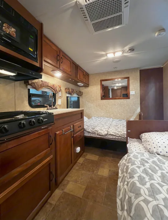 RV kitchen and sleeping area with wood cabinets, stove, microwave, two cozy beds, and tiled floor.. Coachmen Catalina 2012