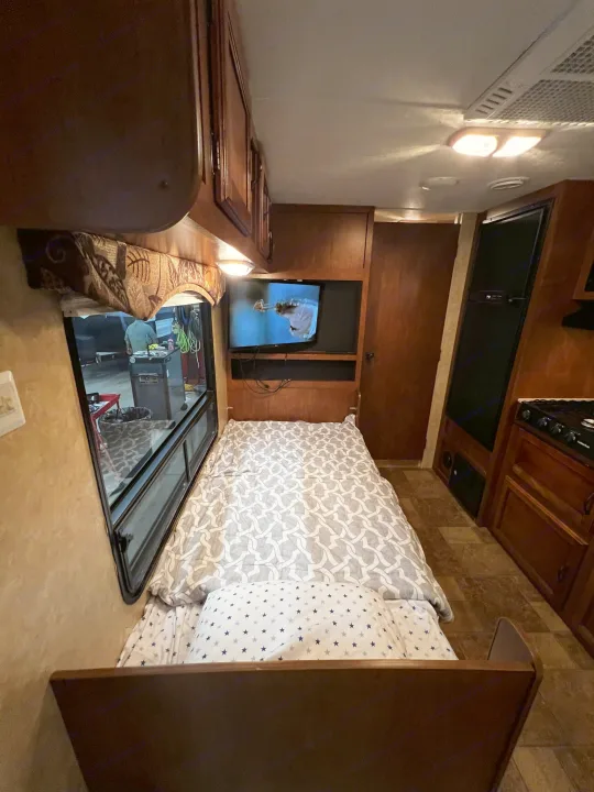 Cozy RV rental interior: bed with TV, compact kitchen, and a window viewing outside. Ideal for comfortable travel.. Coachmen Catalina 2012