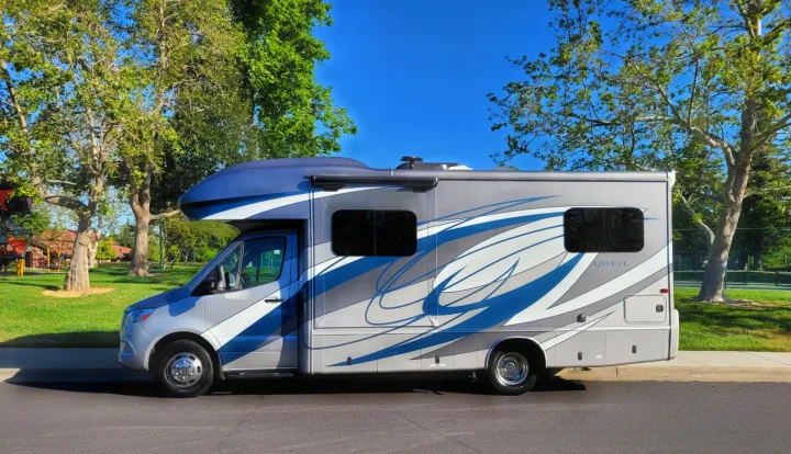 This gray and blue Qwest motorhome is an ideal RV rental. Parked curbside with trees and a clear blue sky.. Entegra Coach Qwest 2020