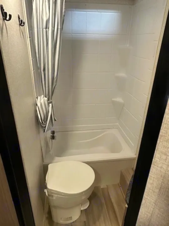 View of a compact RV rental bathroom: white toilet, small tub/shower with striped curtain, and black wall hooks. Practical layout.. Coachmen 2146BHX 2023