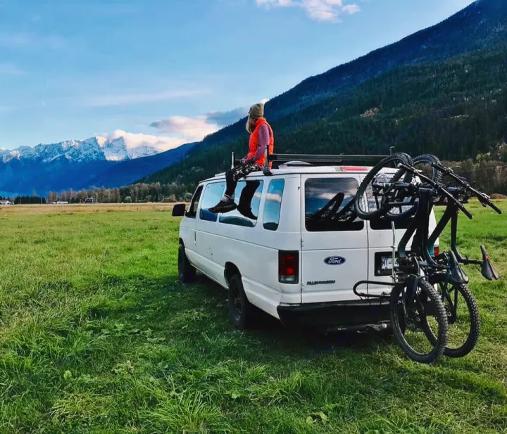 Enjoying mountain views from a white RV rental van with bikes. Person on roof in green field.. Ford Ford E350 1996