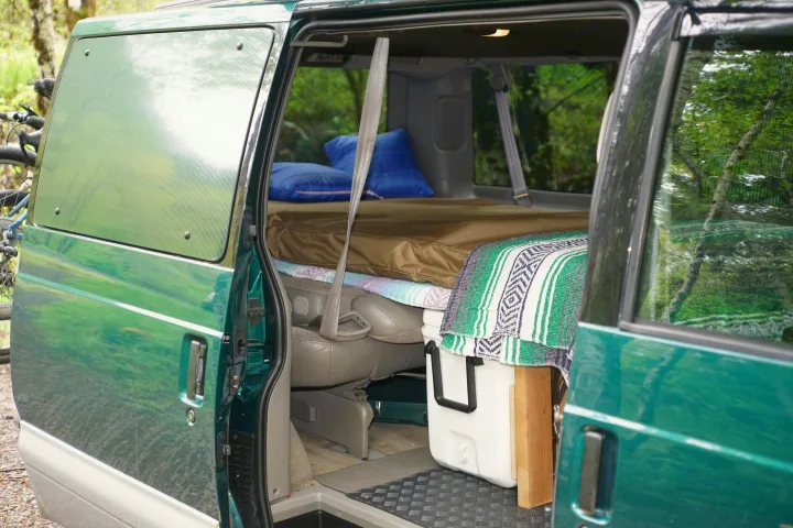 Open side door of a green camper van revealing a bed, pillows, and cooler. An ideal RV rental for outdoor adventures.. Chevrolet Astro Van AWD 1998