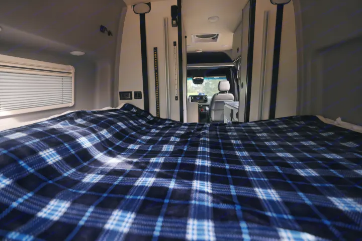 Inside an RV rental: a cozy bed with blue plaid blanket, looking forward to the driver's area. Ready for adventure.. Mercedes Winnebago Revel 2023