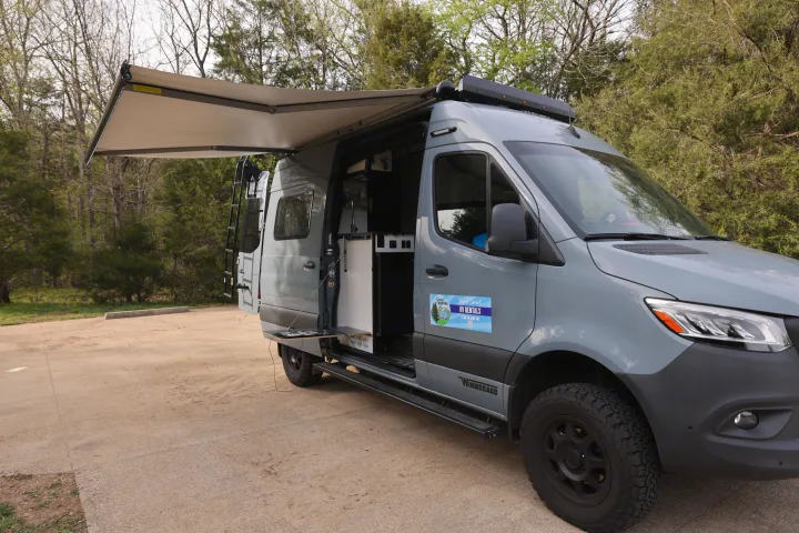 Grey Winnebago RV rental van with an extended awning and open kitchenette door in a park setting.. Mercedes Winnebago Revel 2023