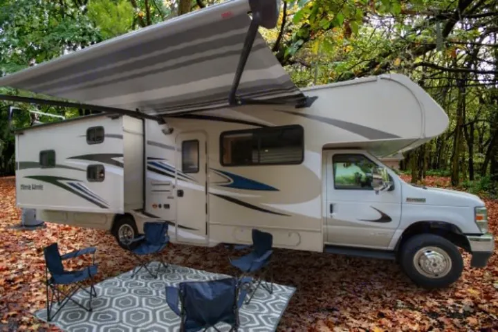 Perfect **RV rental** for camping: Motorhome with extended awning, chairs, and a rug on a leaf-covered ground in a forest.. Winnebago Minnie Winnie 2021