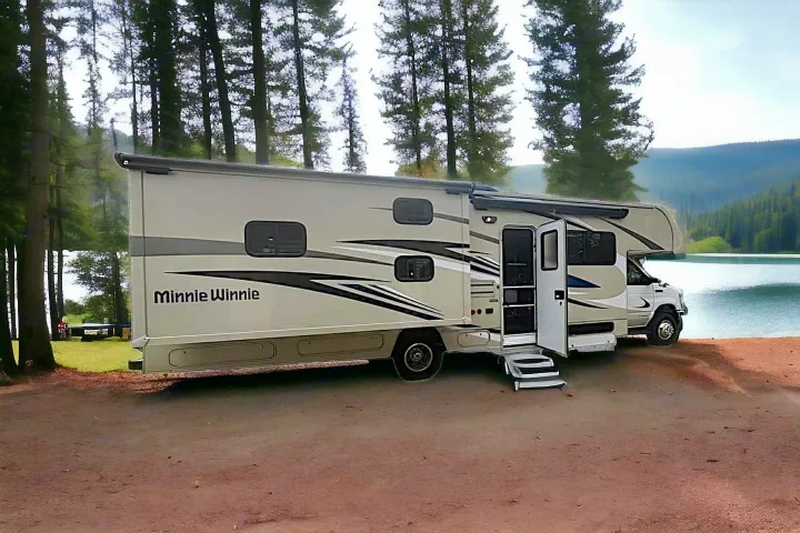 Beige Minnie Winnie RV rental parked by a lake with tall trees, inviting outdoor adventure. Ideal for camping.. Winnebago Minnie Winnie 2021