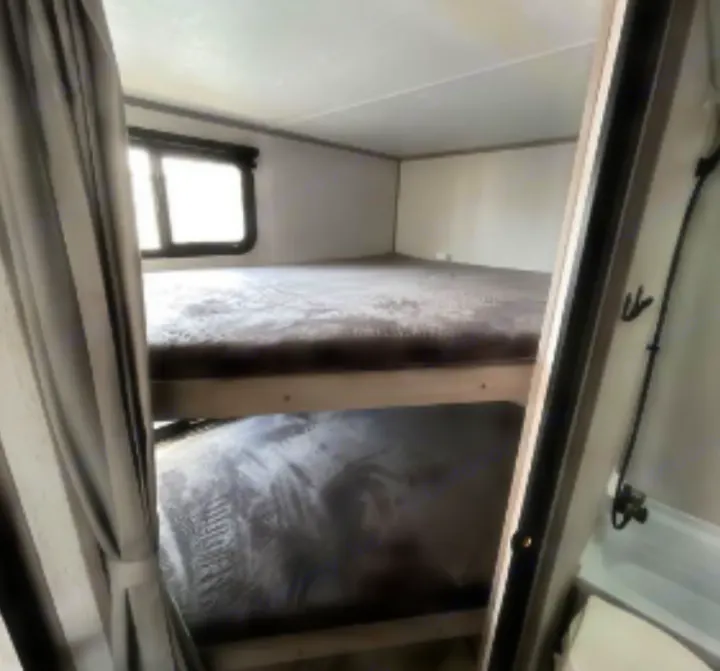 Cozy RV rental interior with bunk beds, soft grey blankets, a window, and a compact bathroom section visible.. Dutchmen Kodiak SE 2023