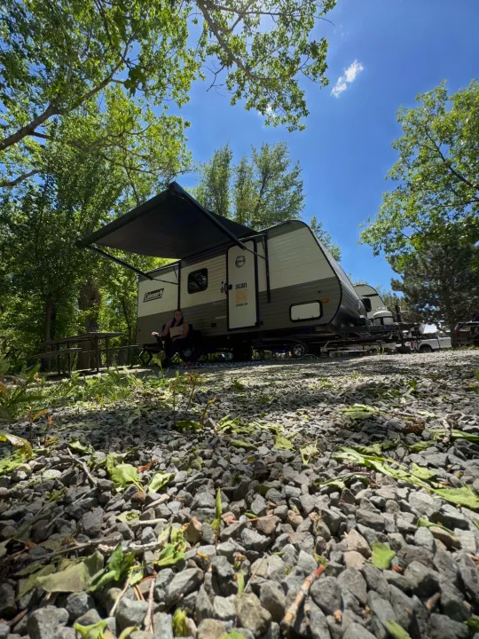 Enjoying the outdoors at a peaceful campsite with an RV rental. A woman relaxes under the awning on a sunny day.. Dutchmen Coleman Lantern LT 2023