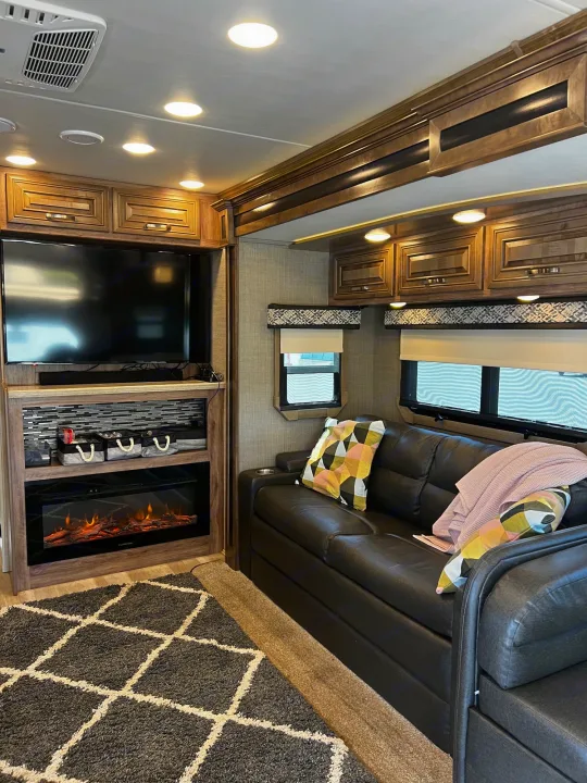 Inside an RV rental: a cozy living area with a black leather couch, electric fireplace, and TV setup.. Jayco Seneca 2021