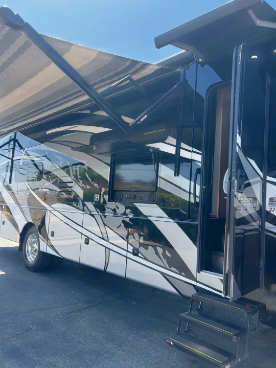 Modern RV rental with an extended awning, outdoor TV setup, and open door showing entry steps. Features stylish black/white graphics.. Jayco Seneca 2021